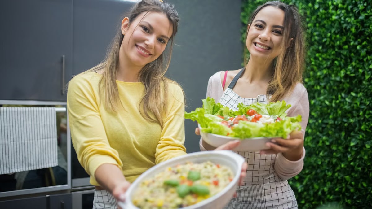 Two women joyfully present fresh vegetable salads in a modern kitchen setting, emphasizing healthy eating.